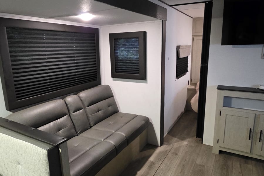 Modern rv rental living area features a gray sofa, built-in dining bench, dark blinds, and wood-look flooring.
