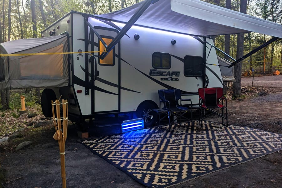 Cozy RV rental campsite with a travel trailer, extended awning, outdoor rug, camping chairs, and ambient lights at dusk.
