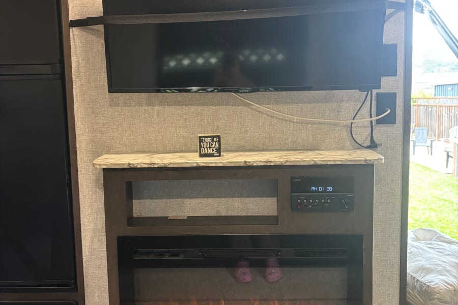 RV rental living space: TV, Furrion stereo, electric fireplace (with purple Crocs!), and a fun "Trust me you can dance" sign.