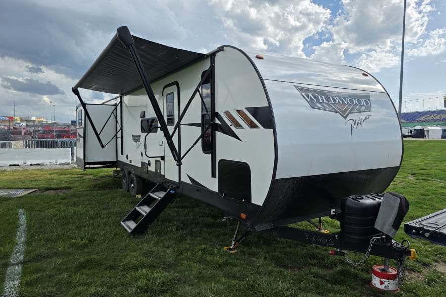 White travel trailer RV rental on grass with awning extended, cloudy sky, and distant stadium seating.