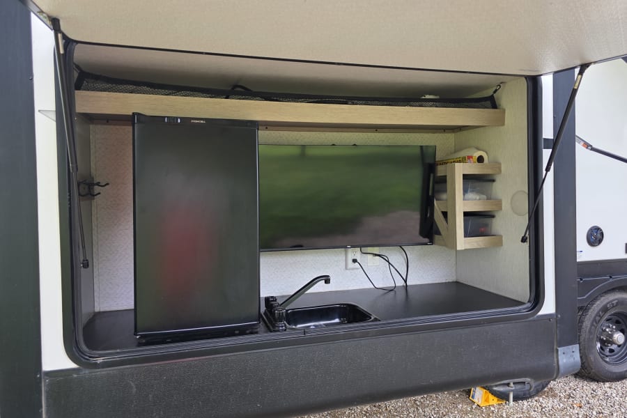 Explore this RV rental's outdoor kitchen: fridge, TV, sink, and shelves with bins, ideal for your next camping trip.