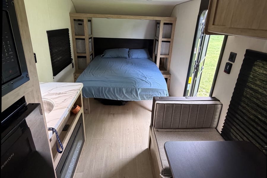 RV rental interior: cozy bedroom with blue bed, kitchen with microwave/sink, and dining booth in a compact living space.