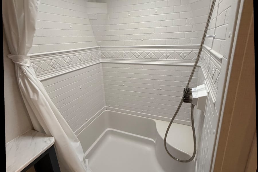 RV rental shower interior: white faux brick walls, decorative trim, shower pan with seat, and tied-back curtain.