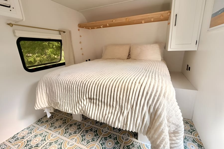 Bright RV rental bedroom featuring a plush bed, stylish patterned floor, window with greenery view, and overhead storage.
