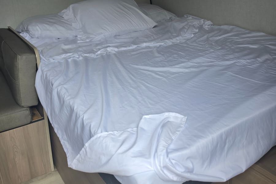 A rumpled white bed with pillows and side seating visible, suggesting a cozy, compact RV rental interior.