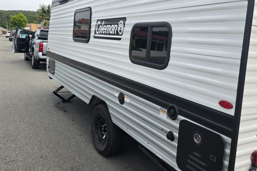 Side view of a striped Coleman travel trailer RV rental, showing windows, utility connections, and wheel, parked next to a truck.