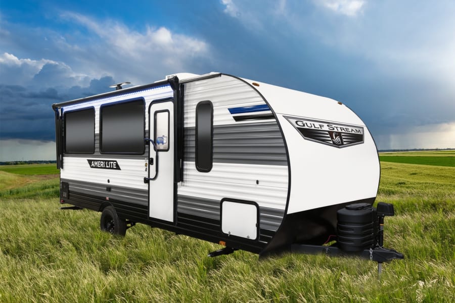 An RV rental: Gulf Stream Ameri Lite travel trailer, white and grey, parked in a vibrant green field under a cloudy sky.