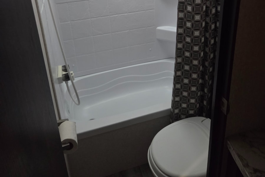 This RV rental bathroom includes a clean white shower/tub with textured wall panels, a patterned curtain, and a toilet.