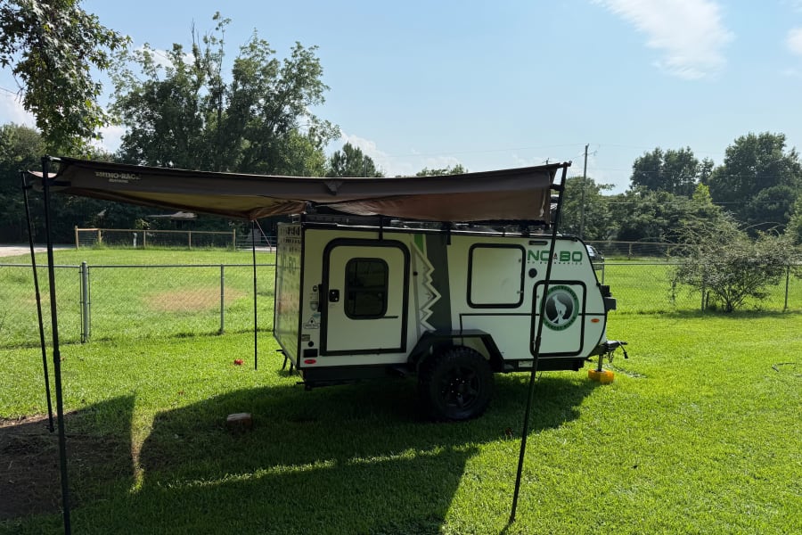 This rv rental, a white NOBO travel trailer with a Rhino-Rack awning, is parked on green grass under a sunny sky.