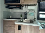 Compact RV rental kitchen with wood-look cabinets, grey backsplash, microwave, and double sink.