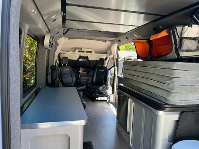 VanHaus ModVan interior.  Seats are swiveled and lower bed is stacked on the right side.  Fridge and extra storage are on the passenger side.