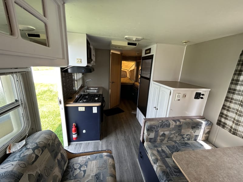 Photo from dining area/lounge (front of camper) with view of kitchenette, cabinetry, refrigerator, bathroom door, and back pop-out queen bed.