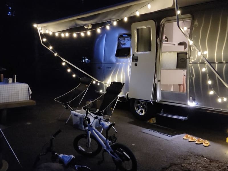 Cozy outdoor setup with Wavago Airstream trailer illuminated by warm string lights. The trailer's awning is extended, creating a welcoming ambiance. Two modern black camping chairs and a small white table are arranged under the lights, perfect for relaxation. In the foreground, a child's bicycle adds a family-friendly touch, while the table and surrounding area suggest an inviting campsite environment. Flip-flops are neatly lined up by the entrance, enhancing the casual, home-away-from-home vibe