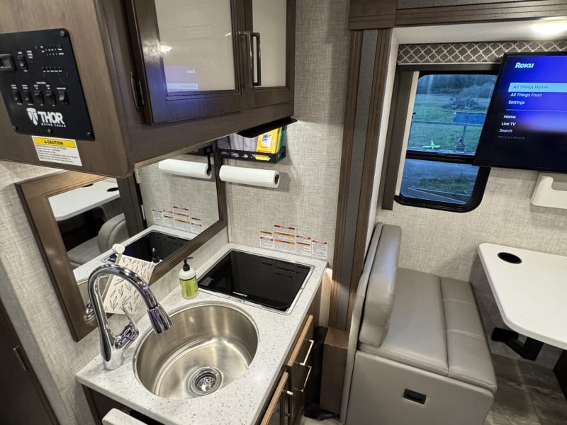 View of sink and stovetop area. Burners have a folding solid cover so that space can be used when burners are not in use. Mounted paper towel holder with storage above. Cabinets above(dishes stored here, as well as knife set, cutting boards) and below(pots and pans here). Drawers with essential kitchen tools stocked(tongs, oven mitt, whisk, measuring cups). 