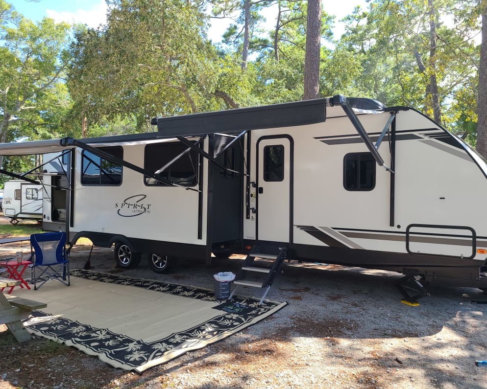 Entrance side with two awnings and dining area slide.  Area rug and camp chairs