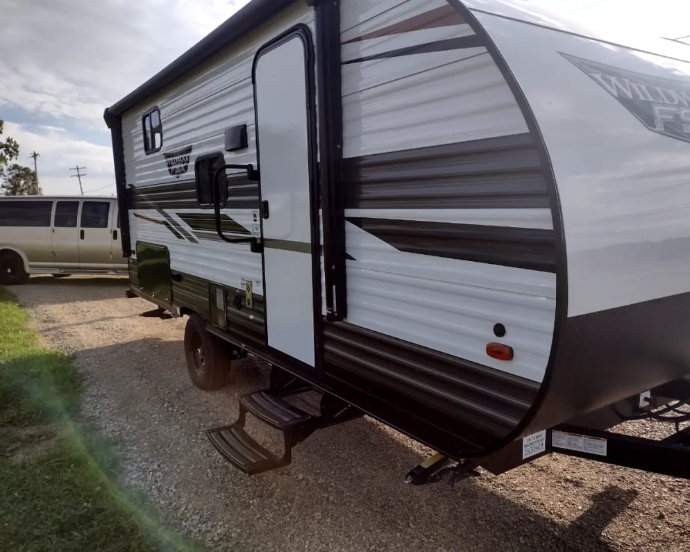 title='Email now about this 2022 Forest River Wildwood FSX Travel Trailer RV for Rent in Fort Wayne, Indiana rental unit!' Wildwood FSX travel trailer rv rental. This white and gray RV with steps is on a gravel lot. A van is in the background.