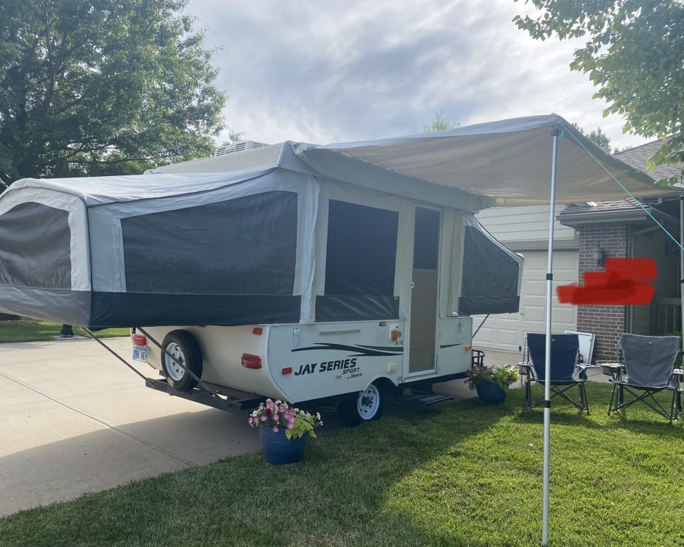 This Jayco pop-up RV rental is fully set up on a driveway with an extended awning over a grassy area with chairs.