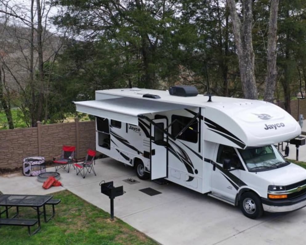 title='Email now about this 2021 Jayco Redhawk SE Class C RV for Rent in Fort Wayne, Indiana rental unit!' A white Jayco rv rental is set up at a spacious campsite, featuring a patio, extended awning, picnic table, and fire pit.