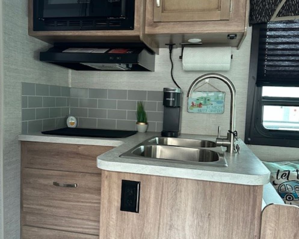 Compact RV rental kitchen with wood-look cabinets, grey backsplash, microwave, and double sink.