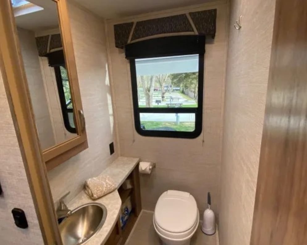 Compact RV rental bathroom with light decor. Features a sink, mirror, toilet, and window showing a campground.