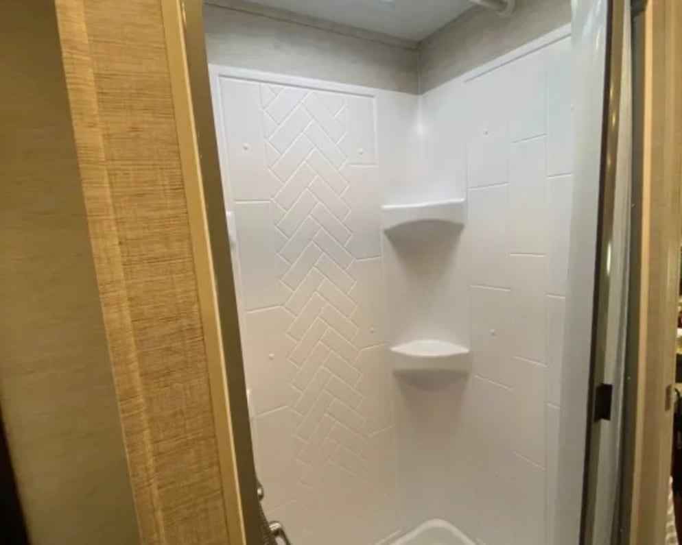 Clean, modern RV rental shower featuring a skylight, white walls with herringbone detail, and a clear door.