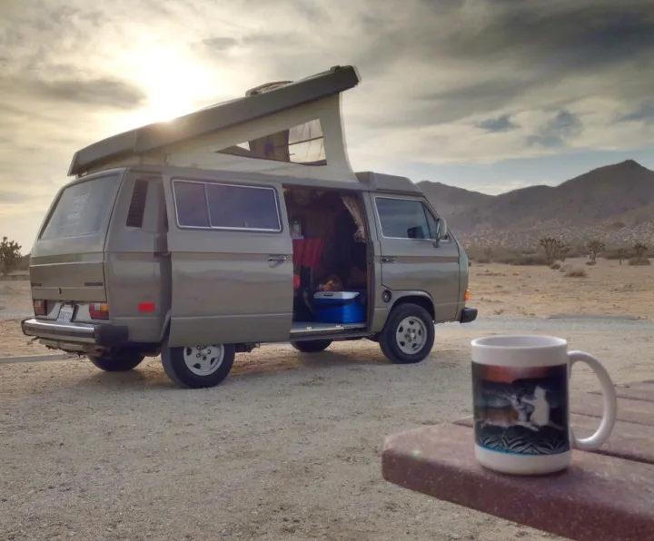 A desert camp scene: a grey pop-top camper van, side door open, with mountains in background. Coffee mug in foreground. Ideal RV rental.. Volkswagen Westfalia 1986