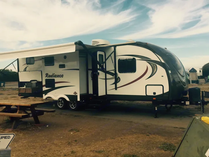 A white Radiance RV rental parked at a dusty campground with an extended awning and a picnic table.. Radiance 28BHIK 2016