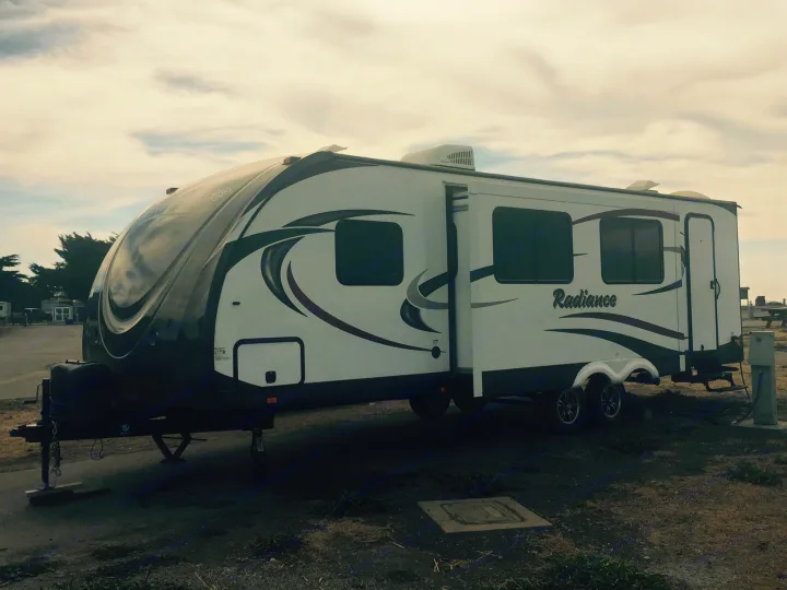White Radiance travel trailer RV rental, parked for a camping trip in a dirt lot under cloudy skies.. Radiance 28BHIK 2016