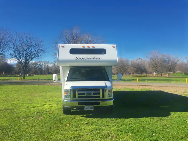 White Sunseeker RV rental parked on a sunny green field with bare trees in the background.. Forest River Sunseeker 2012