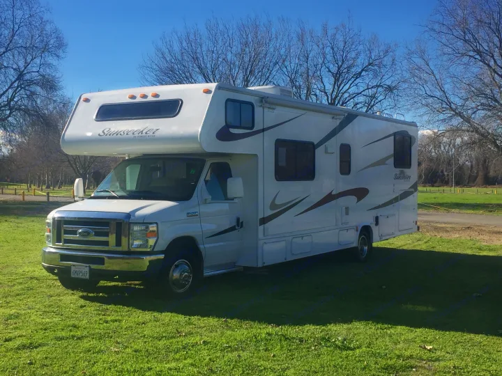 White Sunseeker Class C RV rental parked on sunny green grass with trees. Ideal for outdoor adventures.. Forest River Sunseeker 2012