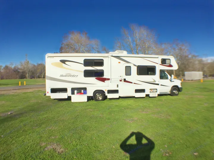 A white Sunseeker RV rental motorhome is parked on bright green grass under a clear blue sky, with its storage compartments open.. Forest River Sunseeker 2012