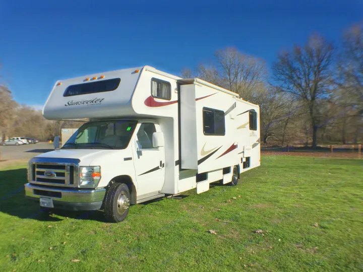 White Sunseeker Class C RV rental with extended slide-out, parked on green grass under a clear blue sky.. Forest River Sunseeker 2012