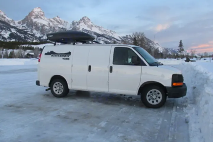 White Basecamper van RV rental with roof box, parked in snowy terrain near majestic mountains, ideal for winter trips.. Chevrolet Express G1500 AWD 2014