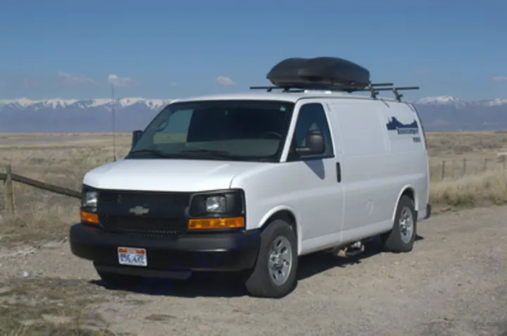 White cargo van with roof rack, ideal for RV rental adventures, parked on a dirt road with snow-capped mountains in the distance.. Chevrolet Express G1500 AWD 2014