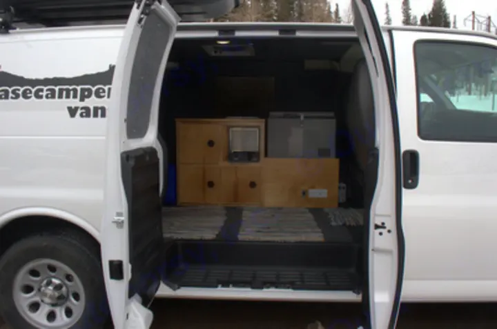 Open doors of an rv rental camper van reveal a practical custom interior with wood storage, fridge, and heater.. Chevrolet Express G1500 AWD 2014