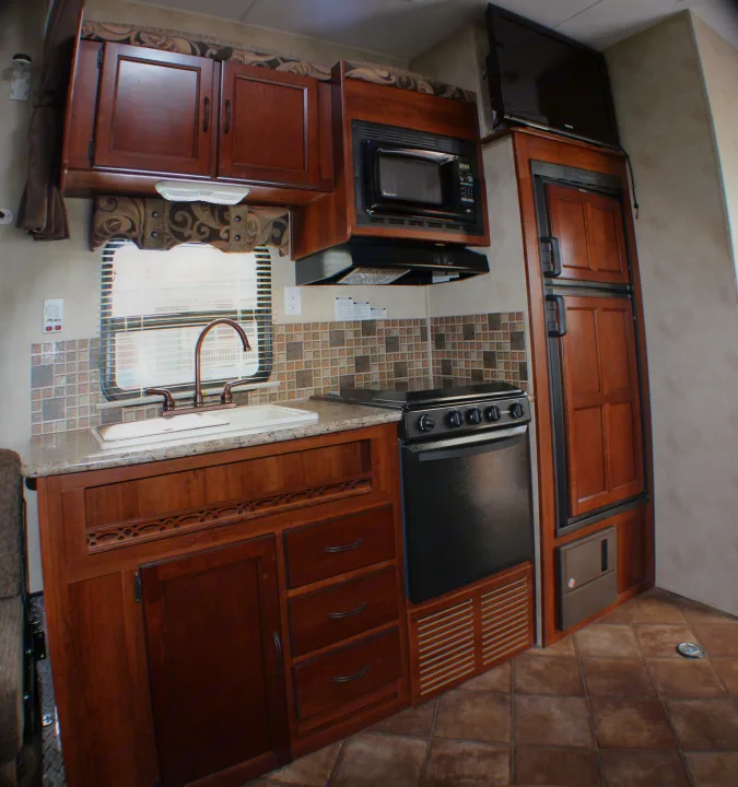 RV rental kitchen: dark wood cabinets, microwave, stove, sink, fridge, TV, tile backsplash & floor fill this compact space.. Palomino Puma Unleashed 2013