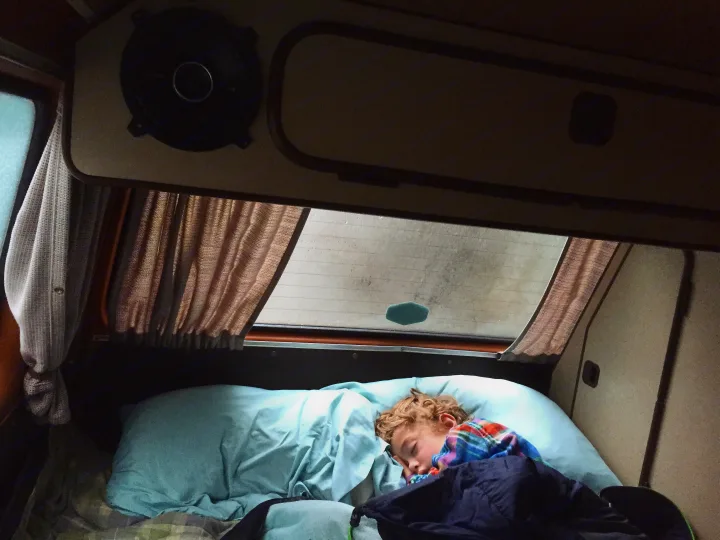A child peacefully sleeps in a cozy RV rental bed, nestled inside a camper van with curtains & speaker visible, perfect for travel.. Volkswagen Vanagon 1985