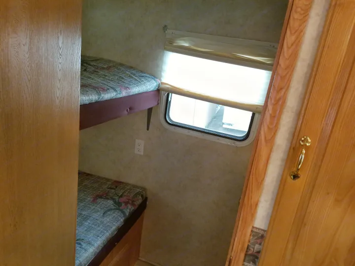 RV rental interior: two stacked bunk beds with blue/red patterned mattresses, a window with light blinds, and a white wall outlet.. Forest River Cedar Creek Silverback Edition 2005
