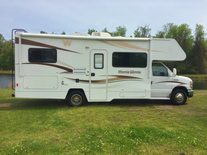 This white Minnie Winnie Class C **RV rental** is parked on green grass by a calm lake with trees, ready for your next adventure.. Winnebago Minnie Winnie 2015