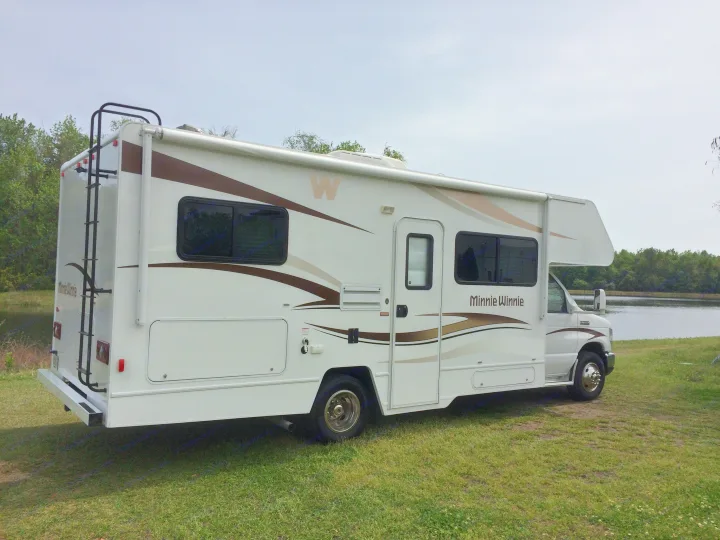 A white and brown Winnebago Minnie Winnie RV, ideal for an rv rental, parked by a lake on green grass.. Winnebago Minnie Winnie 2015