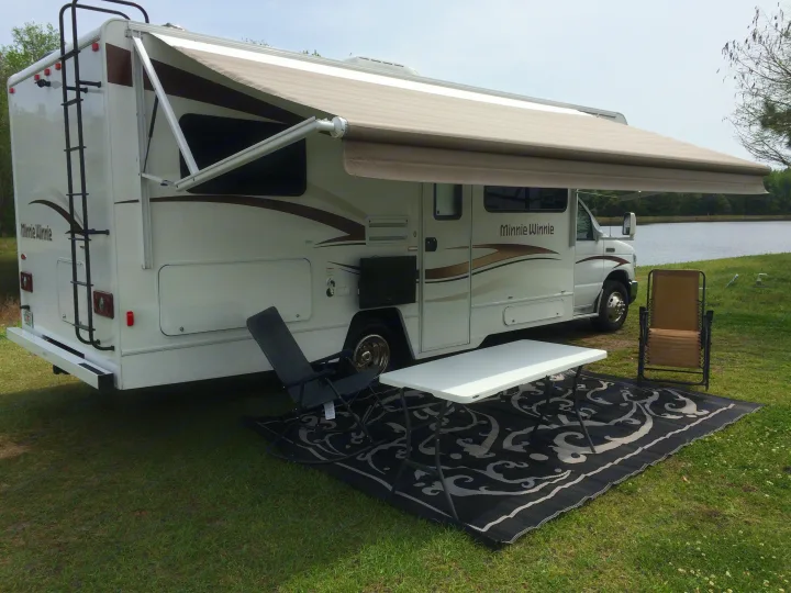 Relaxing RV rental setup: a Minnie Winnie RV with extended awning, outdoor rug, table, and chairs by a serene lake.. Winnebago Minnie Winnie 2015