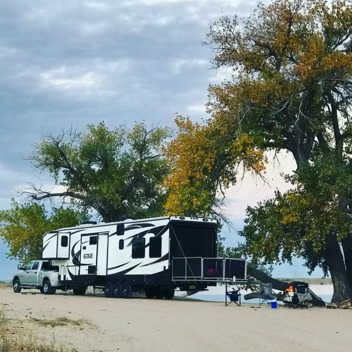 Enjoying an RV rental adventure! A large toy hauler and truck are parked by a lake, with a campfire near autumn trees.. Heartland Edge 2016
