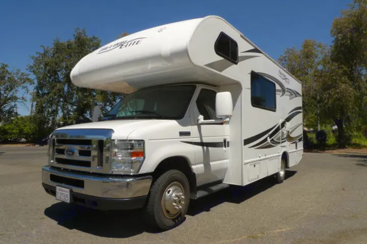 Bright white Class C RV rental, featuring black decals, parked on asphalt with trees under a clear blue sky.. Forest River Freedom 2013