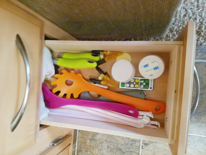 An open RV rental kitchen drawer revealing an orange pasta server, pink spatula, green tongs, remote, and wrapped straws.. Thor Motor Coach Chateau 2011