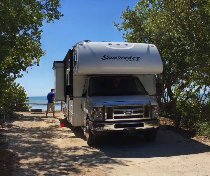 White Sunseeker rv rental parked on a sandy campsite by the water, a man stands enjoying the ocean view.. Forest River Sunseeker 2017