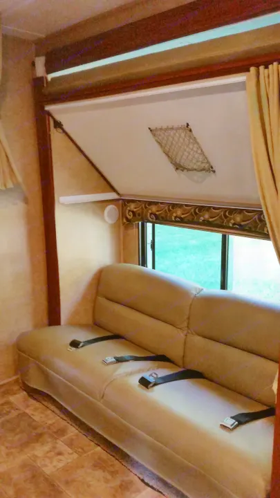 This RV rental interior shows a tan sofa with seatbelts, a large window overlooking grass, and an overhead bed with a mesh net.. Forest River Sunseeker LE 2015