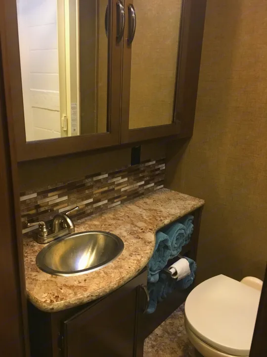 A compact RV rental bathroom featuring a vanity with sink, modern faucet, mirror, toilet, and neatly rolled towels.. Other Other 2018