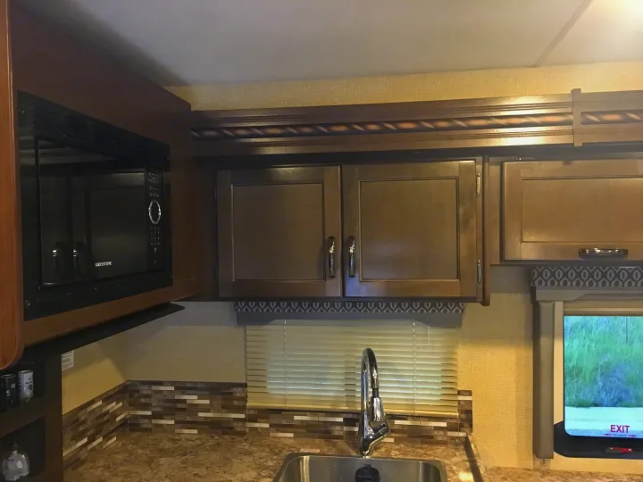 An RV rental kitchen with a Greystone microwave, wood cabinets, and a sink. Notice the tiled backsplash and window.. Other Other 2018