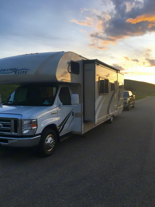 Scenic sunset frames this Freedom Elite Class C **rv rental**, parked on an asphalt road with a truck and rolling hills.. Other Other 2018