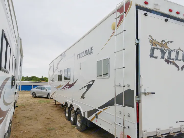 Side view of a white Cyclone Heartland toy hauler RV rental with a ladder & rear ramp, parked in an outdoor lot.. Heartland Cyclone 2009